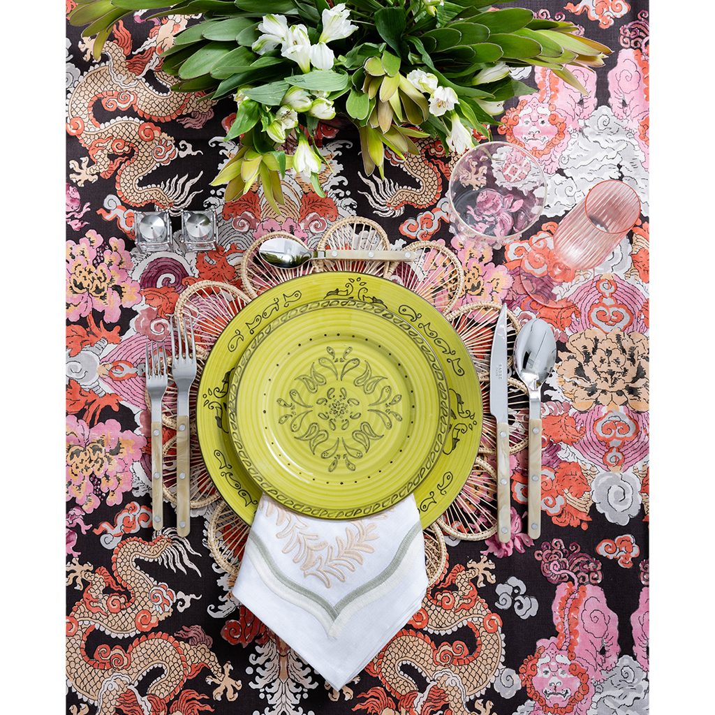 A vibrant tablescape featuring a lime green patterned plate layered over a woven placemat and embroidered white napkin. Colorful dragon-print fabric, polished flatware, pink glassware, and a lush arrangement of white flowers create a bold, playful setting.