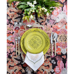 A vibrant tablescape featuring a lime green patterned plate layered over a woven placemat and embroidered white napkin. Colorful dragon-print fabric, polished flatware, pink glassware, and a lush arrangement of white flowers create a bold, playful setting.