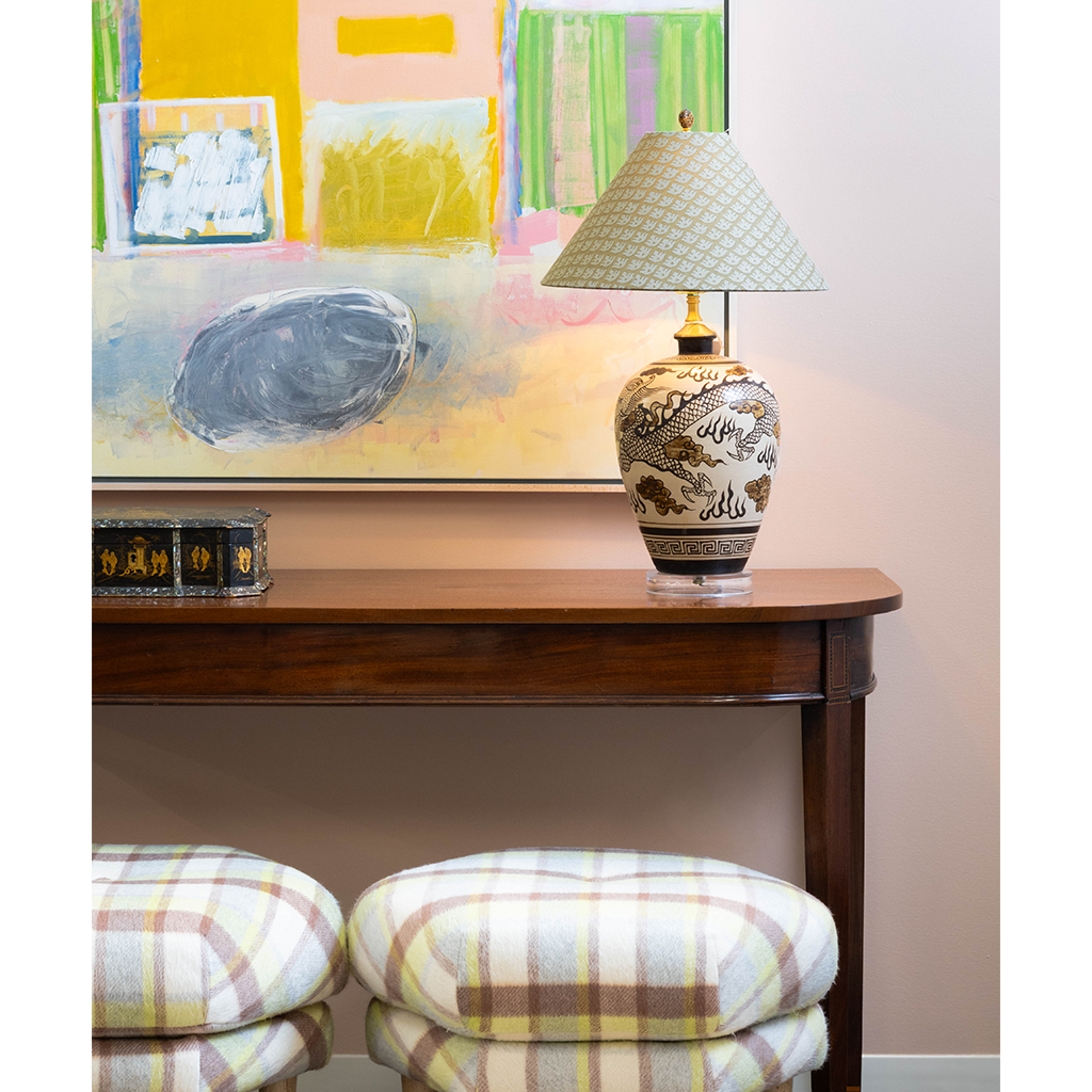 Decorative table lamp with a patterned ceramic base and pale blue geometric shade styled on a dark wood console table. Colorful abstract artwork hangs behind the lamp, while a pair of plaid upholstered ottomans adds soft cream, brown, and yellow tones to the foreground.