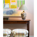 Decorative table lamp with a patterned ceramic base and pale blue geometric shade styled on a dark wood console table. Colorful abstract artwork hangs behind the lamp, while a pair of plaid upholstered ottomans adds soft cream, brown, and yellow tones to the foreground.