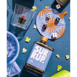 A bottle of SUR RUP. Vanilla flavored malt beverage with a blue label and gold cap, styled on a blue textured surface with a clear gift-themed tumbler, embroidered plate, dice, and small yellow flowers.