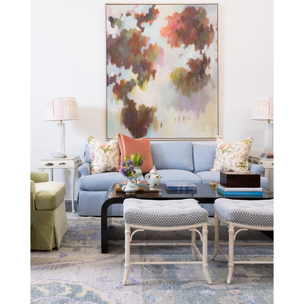 A styled living room featuring a light blue sofa with patterned and solid accent pillows, a black coffee table, two white woven stools, and a large abstract canvas painting on the wall.
