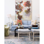 A styled living room featuring a light blue sofa with patterned and solid accent pillows, a black coffee table, two white woven stools, and a large abstract canvas painting on the wall.