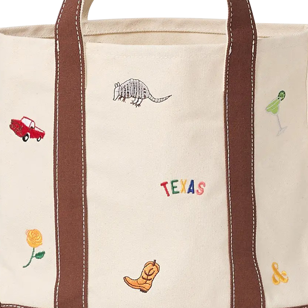 a close up of An off-white canvas tote bag with brown leather handles and base, decorated with small embroidered western-themed motifs including a cowboy boot, an armadillo, a colorful Texas sign, flowers, and a margarita