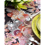 A close-up of a vibrant tablescape featuring a bold multicolored dragon-print tablecloth, woven floral placemat, lime green patterned plates, and pearl-handled flatware. Two small glass candle holders and a lush green floral arrangement add decorative detail to the setting.