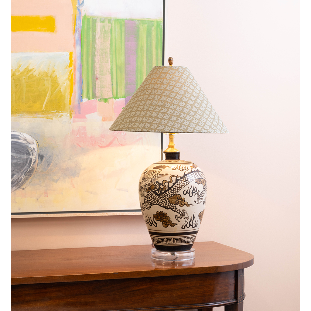 Decorative table lamp featuring a cream ceramic base with an intricate black and brown dragon motif, topped with a pale blue patterned shade. The lamp is styled on a dark wood console table in front of colorful abstract artwork.