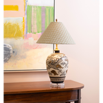 Decorative table lamp featuring a cream ceramic base with an intricate black and brown dragon motif, topped with a pale blue patterned shade. The lamp is styled on a dark wood console table in front of colorful abstract artwork.