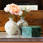 A styled home decor scene featuring two marbled ceramic vases with flowers, set against a wooden dresser background with books.