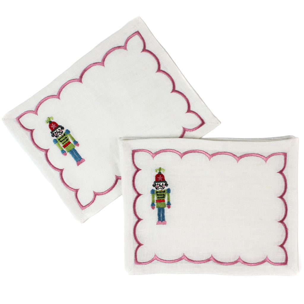 Two white embroidered napkins with pink scalloped borders and a small colorful nutcracker design, shown on a plain white background.