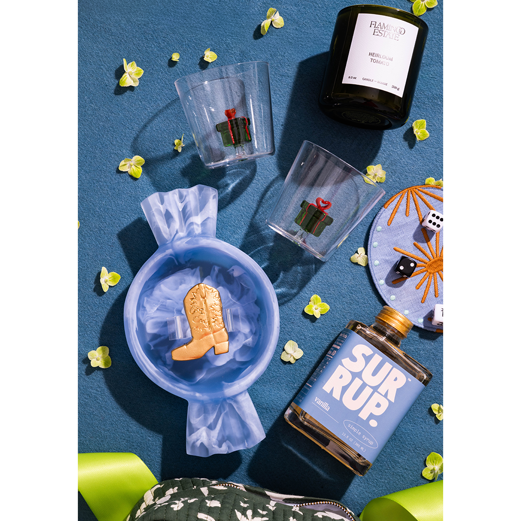 A styled flat-lay product photo featuring a blue glass bottle labeled 'SUR RUP.' and a black candle labeled 'FLAMED EDITION' on a textured blue background, surrounded by small yellow flowers, clear cups with cactus prints, and a blue plate with dice and star details.