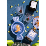 A styled flat-lay product photo featuring a blue glass bottle labeled 'SUR RUP.' and a black candle labeled 'FLAMED EDITION' on a textured blue background, surrounded by small yellow flowers, clear cups with cactus prints, and a blue plate with dice and star details.