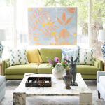 A bright living room featuring a green sofa with patterned and yellow accent pillows, a marble coffee table, and a large abstract wall painting with orange and yellow botanical motifs on a light blue background.