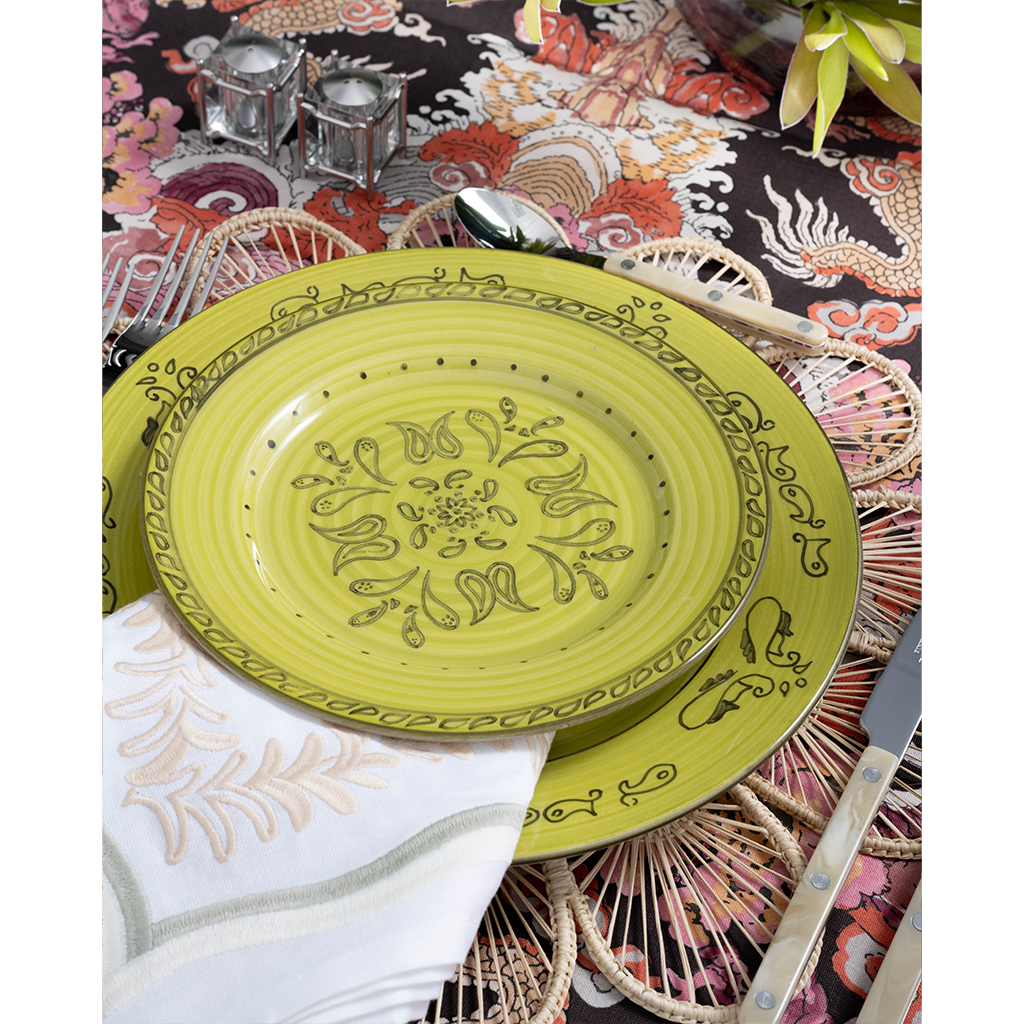 A vibrant place setting featuring a lime green patterned plate layered over a woven floral-shaped placemat and embroidered white napkin. Pearl-handled flatware, pink glassware, and a bold multicolored dragon-print tablecloth complete the playful tablescape.