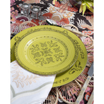 A vibrant place setting featuring a lime green patterned plate layered over a woven floral-shaped placemat and embroidered white napkin. Pearl-handled flatware, pink glassware, and a bold multicolored dragon-print tablecloth complete the playful tablescape.