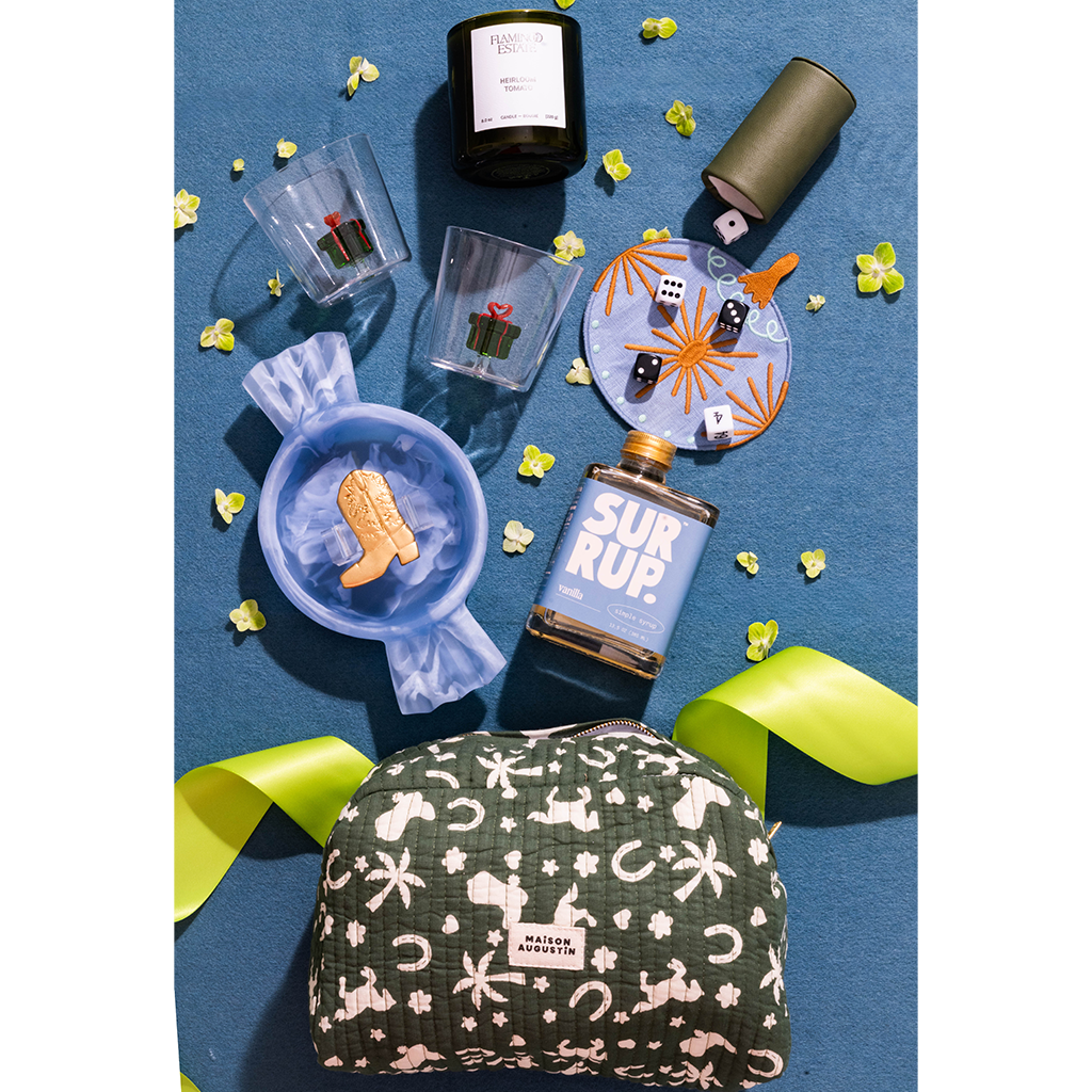 A styled flat-lay of a holiday-themed gift set on a textured blue background, featuring a green and pink bunny print pouch, a blue candy cane candle, a 'SUR RUP.' bottle, and other festive accessories.