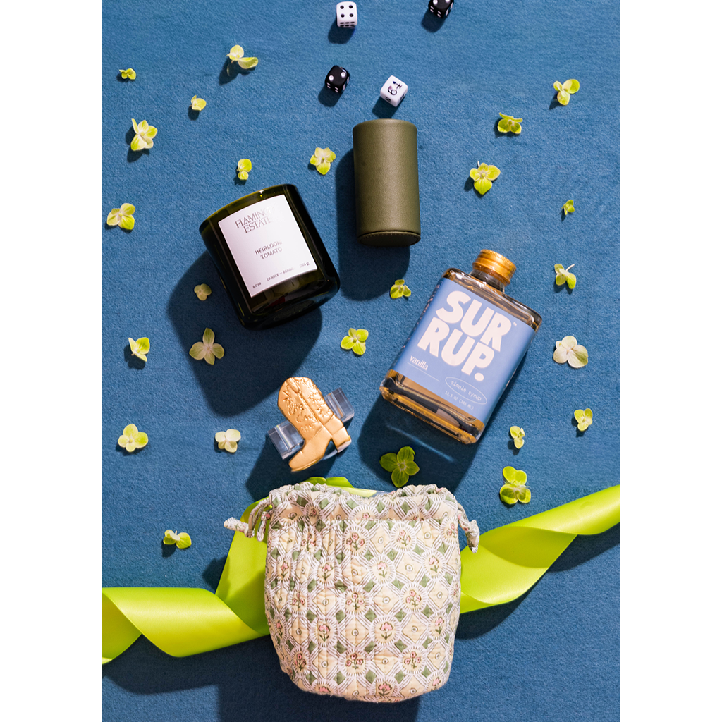A styled flat-lay product photo featuring a floral drawstring pouch, a blue bottle labeled SUR RUP., a black candle jar, a dark green cylindrical container, and a small cork bottle on a textured blue background with scattered yellow flowers and green ribbon.