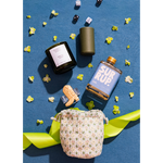 A styled flat-lay product photo featuring a floral drawstring pouch, a blue bottle labeled SUR RUP., a black candle jar, a dark green cylindrical container, and a small cork bottle on a textured blue background with scattered yellow flowers and green ribbon.