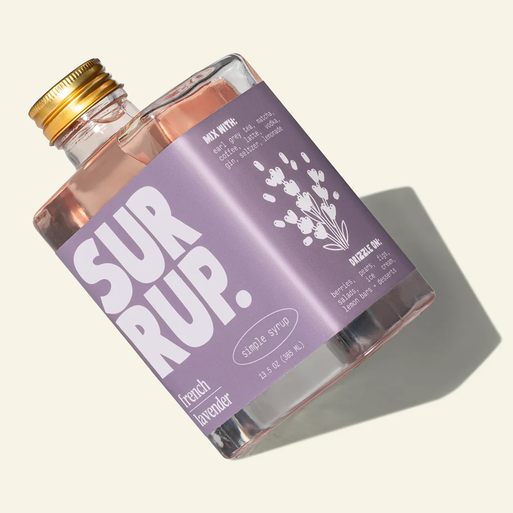 A clear glass bottle of SURUP. French lavender simple syrup with a lavender label and gold cap, shown on a warm off-white background with a soft shadow.