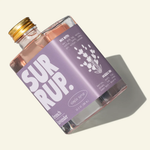 A clear glass bottle of SURUP. French lavender simple syrup with a lavender label and gold cap, shown on a warm off-white background with a soft shadow.