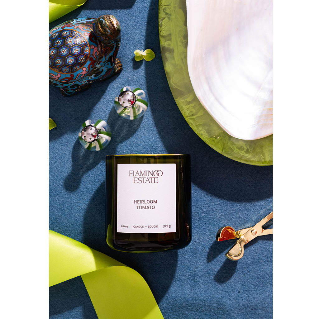A Flamingo Estate 'Herbloom Tomato' scented candle in a dark glass jar, styled in a flat-lay arrangement on a textured blue surface with decorative green leaves, a patterned figurine, and a white fabric piece.