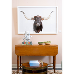 A warm interior vignette featuring a wooden drop-leaf console table styled with decorative bowls, stacked books, and a sculptural accent. A large framed longhorn portrait hangs above, while a round wood and brass coffee table with books sits in the foreground.