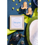 A framed print with the words 'blue print' in blue text on a white background, styled in a flat lay with small cowboy boot-shaped clips, green ribbon, a decorative peacock figurine, and a white plate on a dark blue surface.