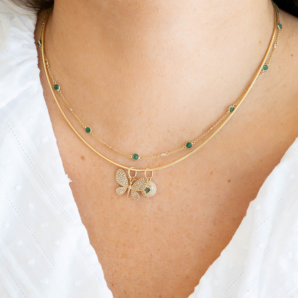 A close-up of a model wearing a double-layered gold necklace. The necklace features a delicate chain with small green round stones and a central butterfly charm encrusted with clear crystals, with a matching round medallion pendant.