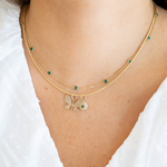 A close-up of a model wearing a double-layered gold necklace. The necklace features a delicate chain with small green round stones and a central butterfly charm encrusted with clear crystals, with a matching round medallion pendant.