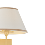 Close-up of a table lamp with a tapered off-white fabric shade trimmed in beige, mounted on a brushed gold metal stand against a white background.