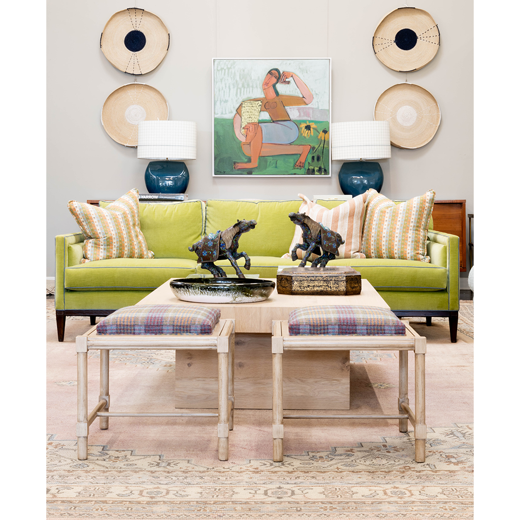 A colorful living room featuring a lime green upholstered sofa layered with patterned pillows, flanked by blue ceramic lamps and woven wall accents. A light wood cocktail table with decorative horse sculptures sits behind two plaid-cushioned stools, while colorful artwork and a patterned rug complete the space.