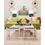 A colorful living room featuring a lime green upholstered sofa layered with patterned pillows, flanked by blue ceramic lamps and woven wall accents. A light wood cocktail table with decorative horse sculptures sits behind two plaid-cushioned stools, while colorful artwork and a patterned rug complete the space.