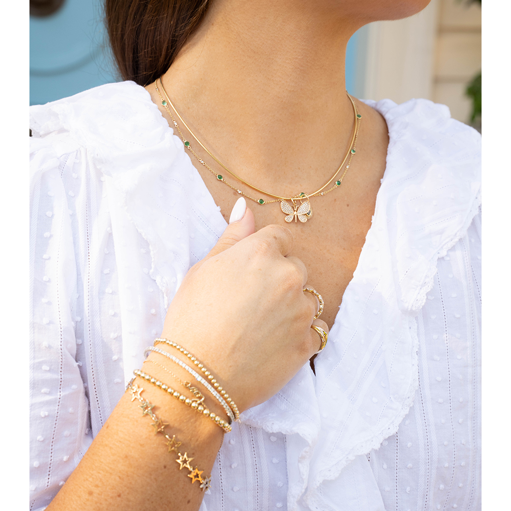 A close-up lifestyle shot of a woman wearing a layered gold necklace with a butterfly pendant and green gemstone accents, along with matching gold bracelets and rings, paired with a white ruffled blouse.