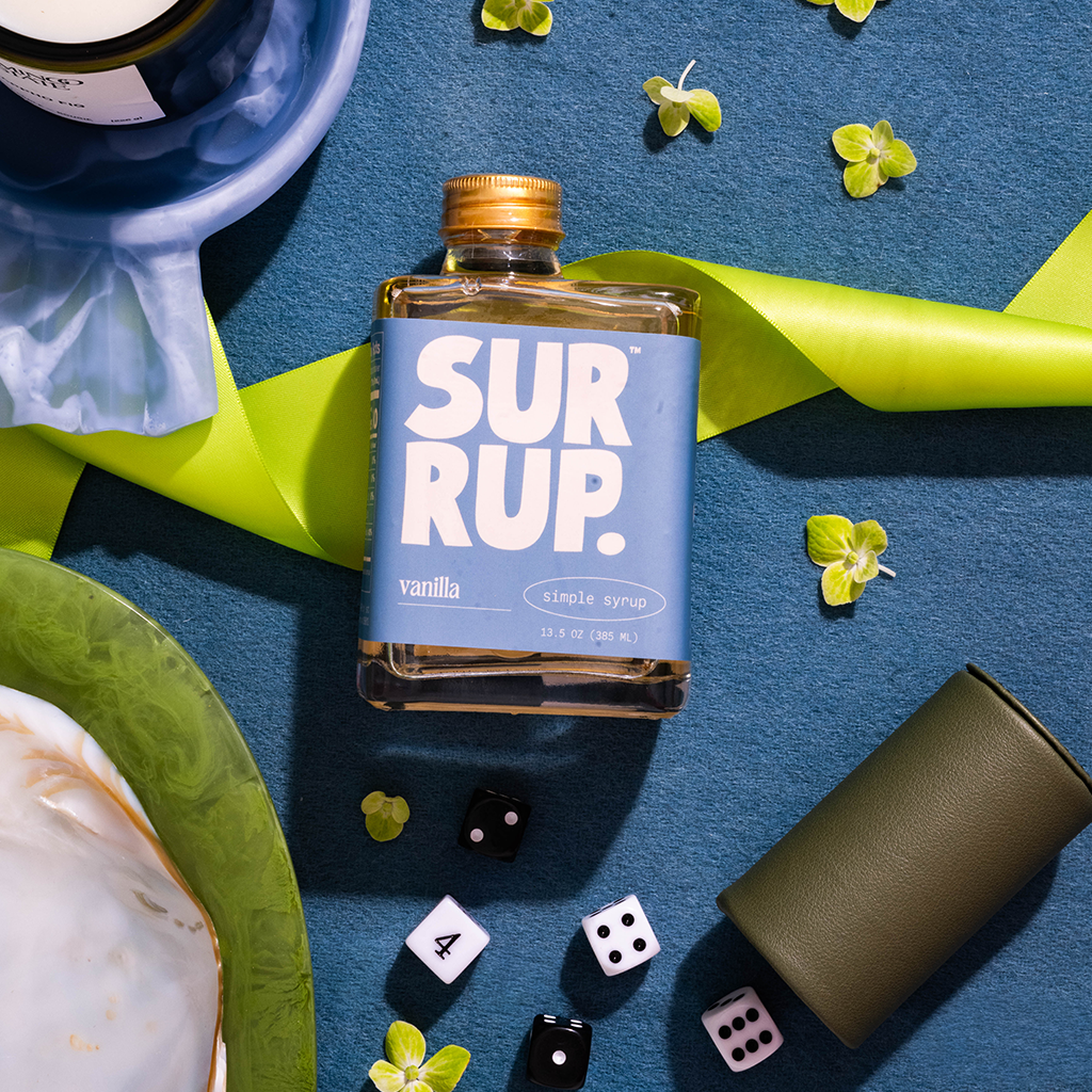 A bottle of SURUP vanilla simple syrup centered on a textured blue surface, surrounded by styled props including a green ribbon, small yellow flowers, dice, a cup, and a plate.