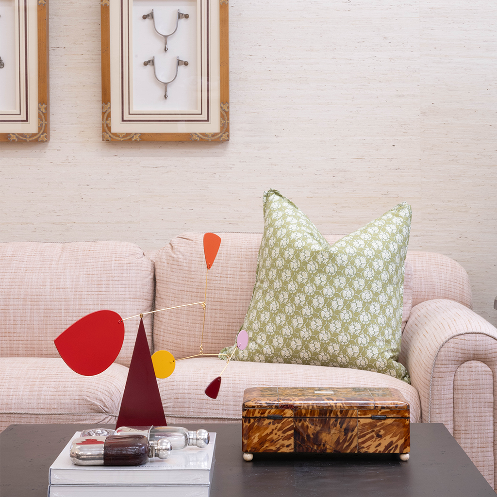 A blush pink upholstered sofa with a green floral throw pillow sits behind a dark cocktail table styled with a colorful sculptural mobile, stacked books, decorative bottles, and a tortoiseshell box. Framed artwork and textured neutral walls complete the layered living room vignette.