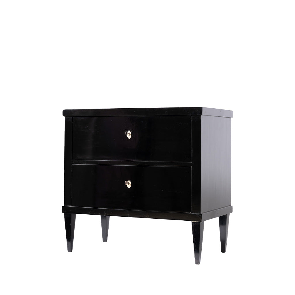 A three-quarter view of an antique ebonized two-drawer chest with a glossy black finish features clean, rectilinear lines, tapered legs, and ivory-colored shield-shaped keyhole escutcheons. The minimalist design and elegant proportions reflect the refined simplicity of the Biedermeier style.