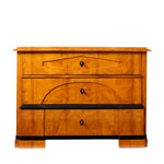 Front view of an antique Biedermeier chest of drawers with a warm honey-colored wood veneer, featuring three graduated drawers with geometric veneer detailing and black shield-shaped keyhole escutcheons. Contrasting ebonized trim, stepped block feet, and clean architectural lines emphasize the piece's refined craftsmanship and elegant design.