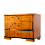 A three-quarter view of an antique Biedermeier chest of drawers with a warm honey-colored wood veneer features three graduated drawers with geometric veneer detailing, black shield-shaped keyhole escutcheons, and contrasting ebonized trim. The clean lines, stepped block feet, and striking wood grain exemplify the refined elegance of the Biedermeier style.