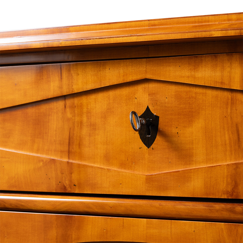 A close-up of an antique Biedermeier chest of drawers highlights the warm honey-colored wood veneer with striking geometric veneer patterns across the drawer front. A black shield-shaped keyhole escutcheon with its original key adds a distinctive decorative detail.