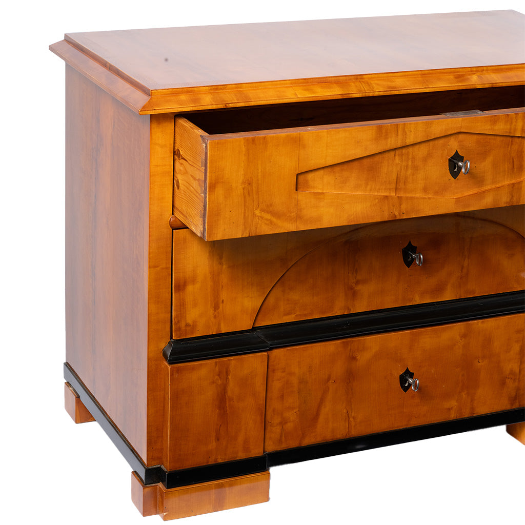 A close-up, three-quarter view of an antique Biedermeier chest of drawers shows the top drawer partially open, revealing its dovetail construction and solid wood interior. The warm honey-colored wood veneer, ebonized trim, and geometric veneer details highlight the piece's fine craftsmanship.