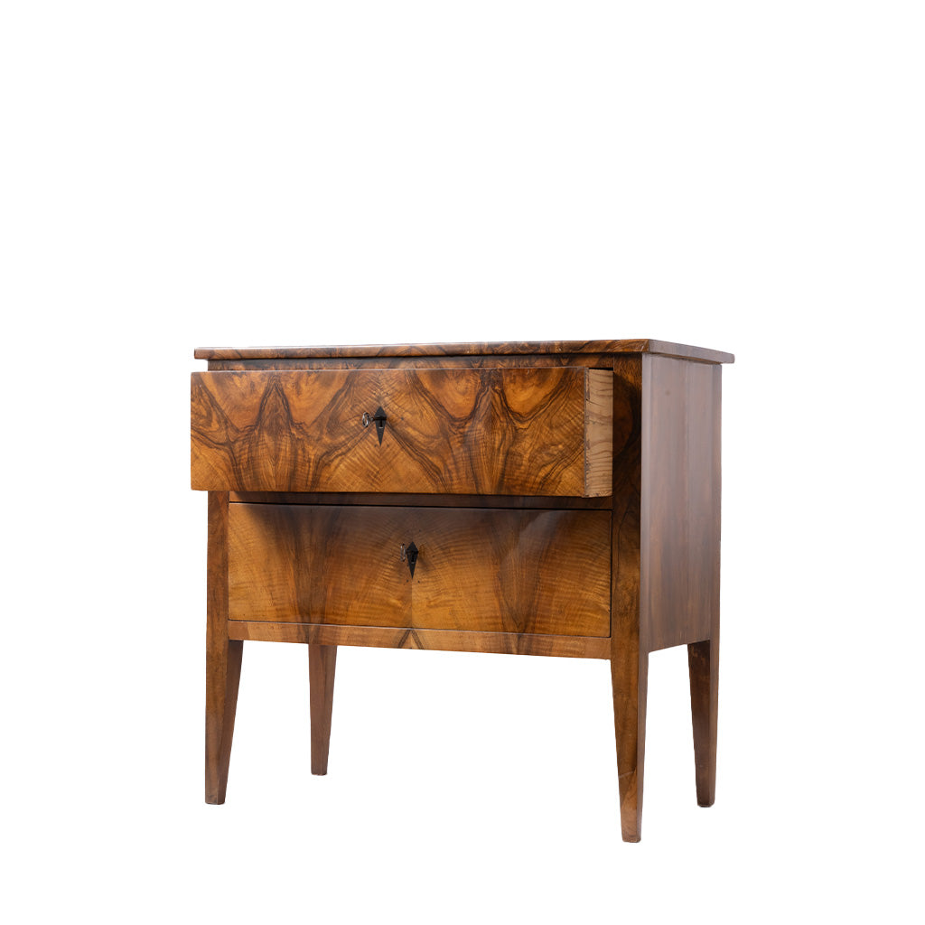 A three-quarter view of an antique two-drawer chest with striking bookmatched walnut veneer showcases the dramatic wood grain across the drawer fronts. The piece features simple keyhole escutcheons, tapered legs, and clean lines characteristic of the elegant Biedermeier style.