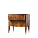 A three-quarter view of an antique two-drawer chest with striking bookmatched walnut veneer showcases the dramatic wood grain across the drawer fronts. The piece features simple keyhole escutcheons, tapered legs, and clean lines characteristic of the elegant Biedermeier style.