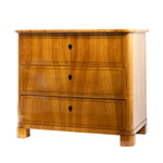 A front view of an antique Biedermeier chest of drawers in a warm honey-colored wood veneer features three graduated drawers with keyholes and delicate string inlay. Rounded front corners, clean lines, and block feet emphasize the piece's refined, understated design.