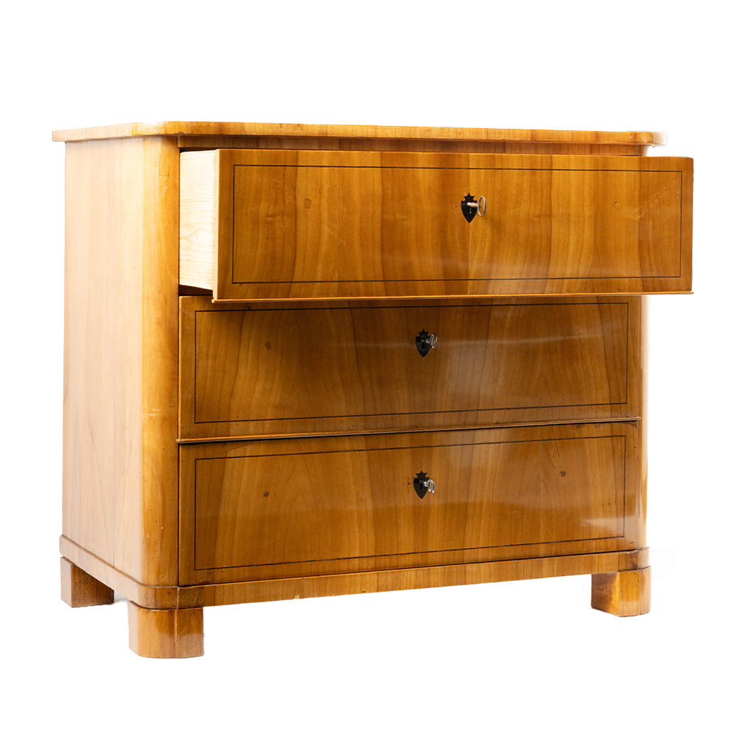 A three-quarter view of an antique chest of drawers with a warm honey-colored wood veneer features three graduated drawers with keyholes and subtle string inlay. The clean lines, rounded front corners, and block feet reflect the elegant simplicity of the Biedermeier style.