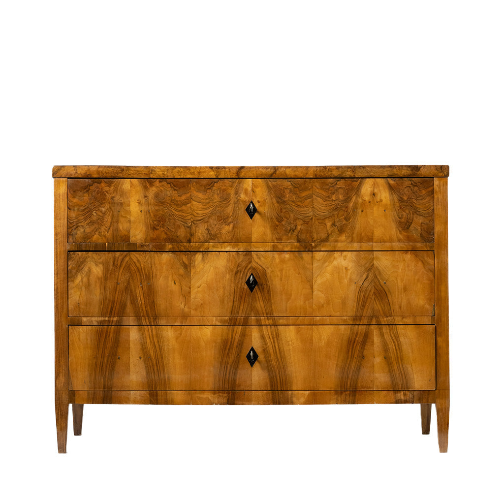 A full frontal view of an antique Biedermeier chest of drawers revealing dovetail joinery and a solid wood interior. The richly figured walnut veneer, dramatic bookmatched grain, and simple black diamond-shaped keyhole escutcheons highlight the piece's fine craftsmanship.