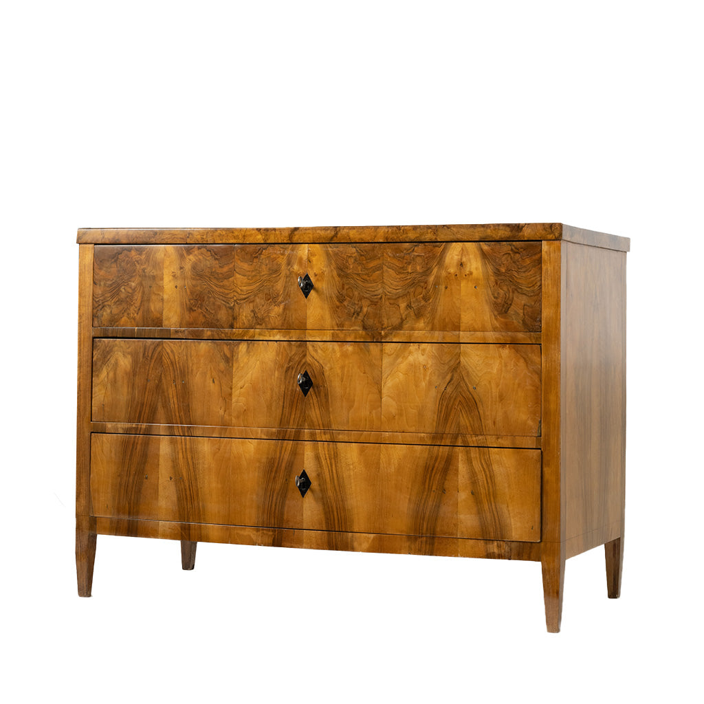 A front view of an antique Biedermeier chest of drawers revealing dovetail joinery and a solid wood interior. The richly figured walnut veneer, dramatic bookmatched grain, and simple black diamond-shaped keyhole escutcheons highlight the piece's fine craftsmanship.