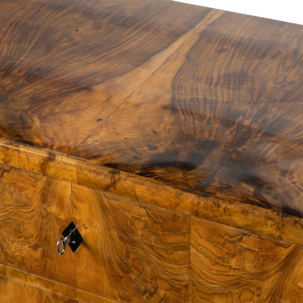 Close-up of a wooden furniture piece showing richly figured burl wood grain in warm golden-brown and dark brown tones, with a small black metal lock on the front.