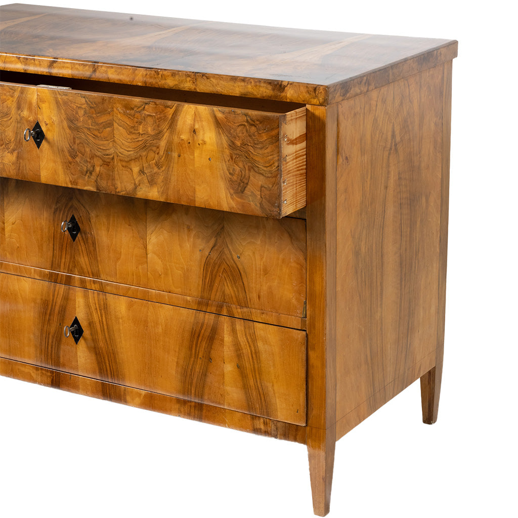 A close-up, three-quarter view of an antique Biedermeier chest of drawers shows the top drawer partially open, revealing dovetail joinery and a solid wood interior. The richly figured walnut veneer, dramatic bookmatched grain, and simple black diamond-shaped keyhole escutcheons highlight the piece's fine craftsmanship.