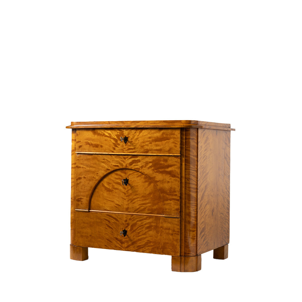 A three-quarter view of an antique Biedermeier chest of drawers shows the top drawer partially open, revealing dovetail joinery and a solid wood interior. The richly figured golden burl wood veneer, arched veneer detailing across the lower drawers, and block feet highlight the piece's exceptional craftsmanship and elegant design.