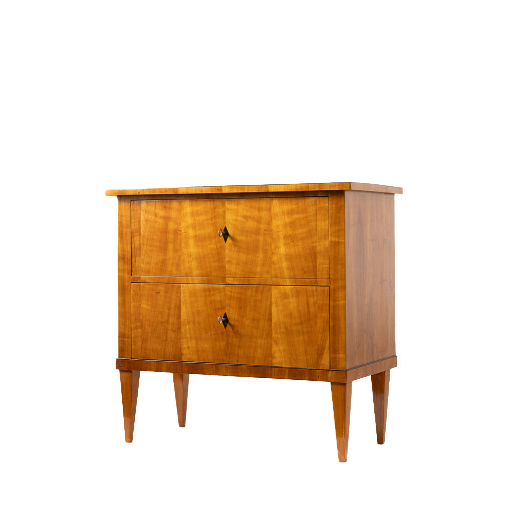 A three-quarter view of an antique Biedermeier two-drawer chest features a warm honey-colored wood veneer with beautifully matched figured grain across the drawer fronts. Simple diamond-shaped pulls, tapered legs, and clean, elegant lines highlight the piece's refined craftsmanship and timeless design.