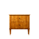 A three-quarter view of an antique Biedermeier two-drawer chest, revealing dovetail joinery and a solid wood interior. The chest features a warm honey-colored wood veneer, simple round pulls, tapered legs, and clean lines that highlight its refined craftsmanship.